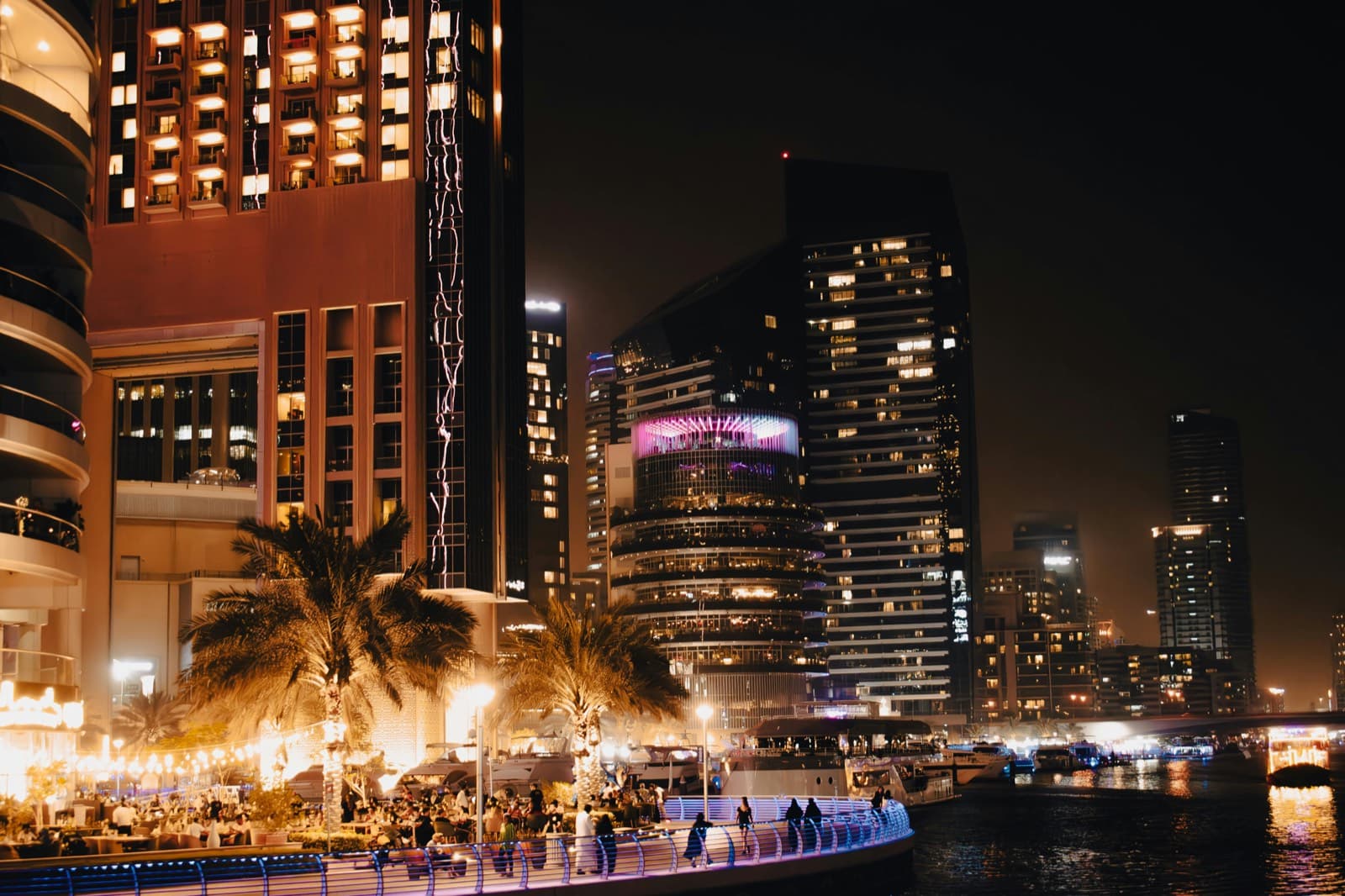 Večerní promenáda Dubai Marina s osvětlenými mrakodrapy, palmami a lidmi na procházce